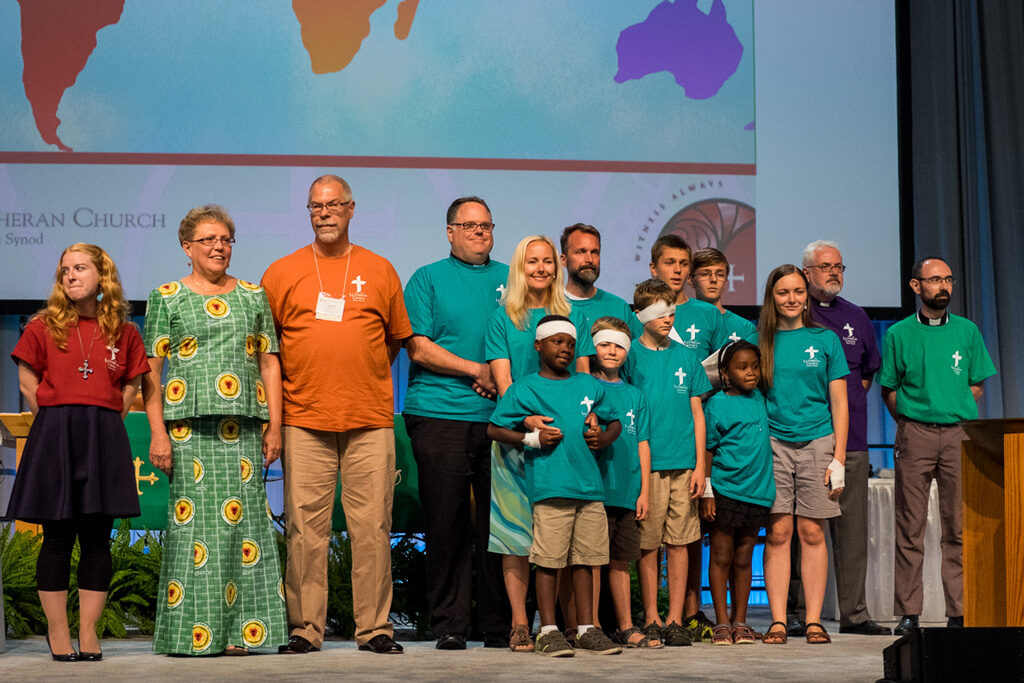 Six career missionaries, with their spouses and families, stand on the convention stage for a special recognition of their service on Monday, July 11, in Milwaukee. (LCMS/Michael Schuermann)
